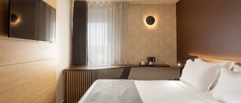 Une chambre d’hôtel moderne dotée d’un grand lit, d’un téléviseur à écran plat fixé au mur, d’une table de chevet avec une lampe et d’une fenêtre ornée de rideaux légers.