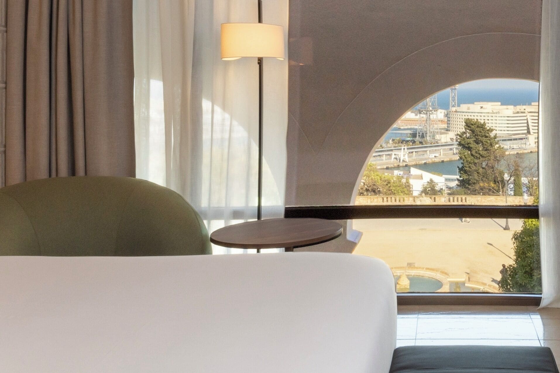 Une chambre d’hôtel avec vue sur la ville, un fauteuil vert, une petite table ronde et une lampe.