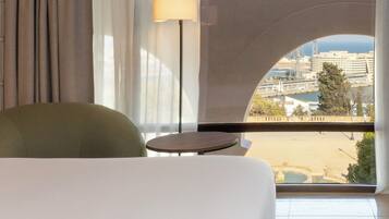 Une chambre d’hôtel avec vue sur la ville, un fauteuil vert, une petite table ronde et une lampe.