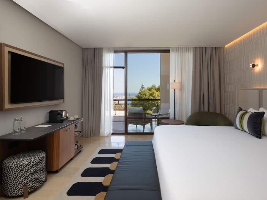Ein Hotelzimmer mit einem großen Bett, einem Flachbildfernseher, einem Schreibtisch mit Kaffeemaschine, einem kleinen Tisch und einem Balkon mit Aussicht.
