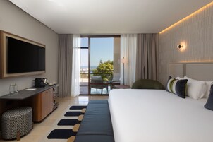 Ein Hotelzimmer mit einem großen Bett, einem Flachbildfernseher, einem Schreibtisch mit Kaffeemaschine, einem kleinen Tisch und einem Balkon mit Aussicht.