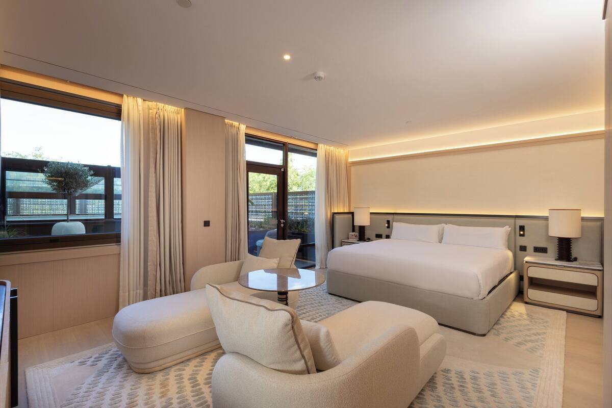 Ein modernes Hotelzimmer mit einem großen Bett, zwei Sesseln, einem kleinen runden Tisch und Blick ins Freie durch ein großes Fenster.