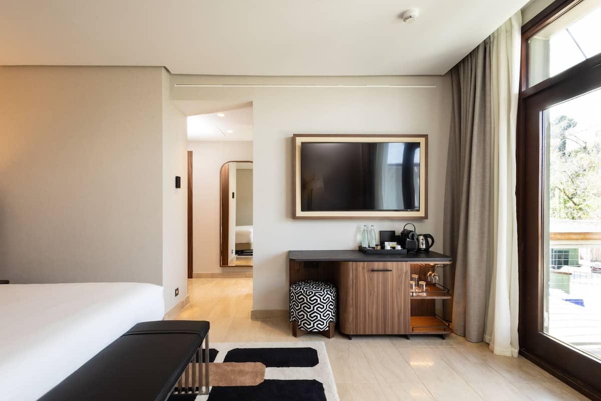 Ein modernes Hotelzimmer mit einem großen Bett, einem Schreibtisch, einem Flachbildfernseher, einer Küchenzeile und einem großen Fenster mit Vorhängen.