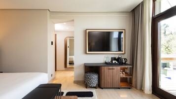 A modern hotel room with a large bed, a desk with a flat-screen TV, a kitchenette, and a large window with curtains.