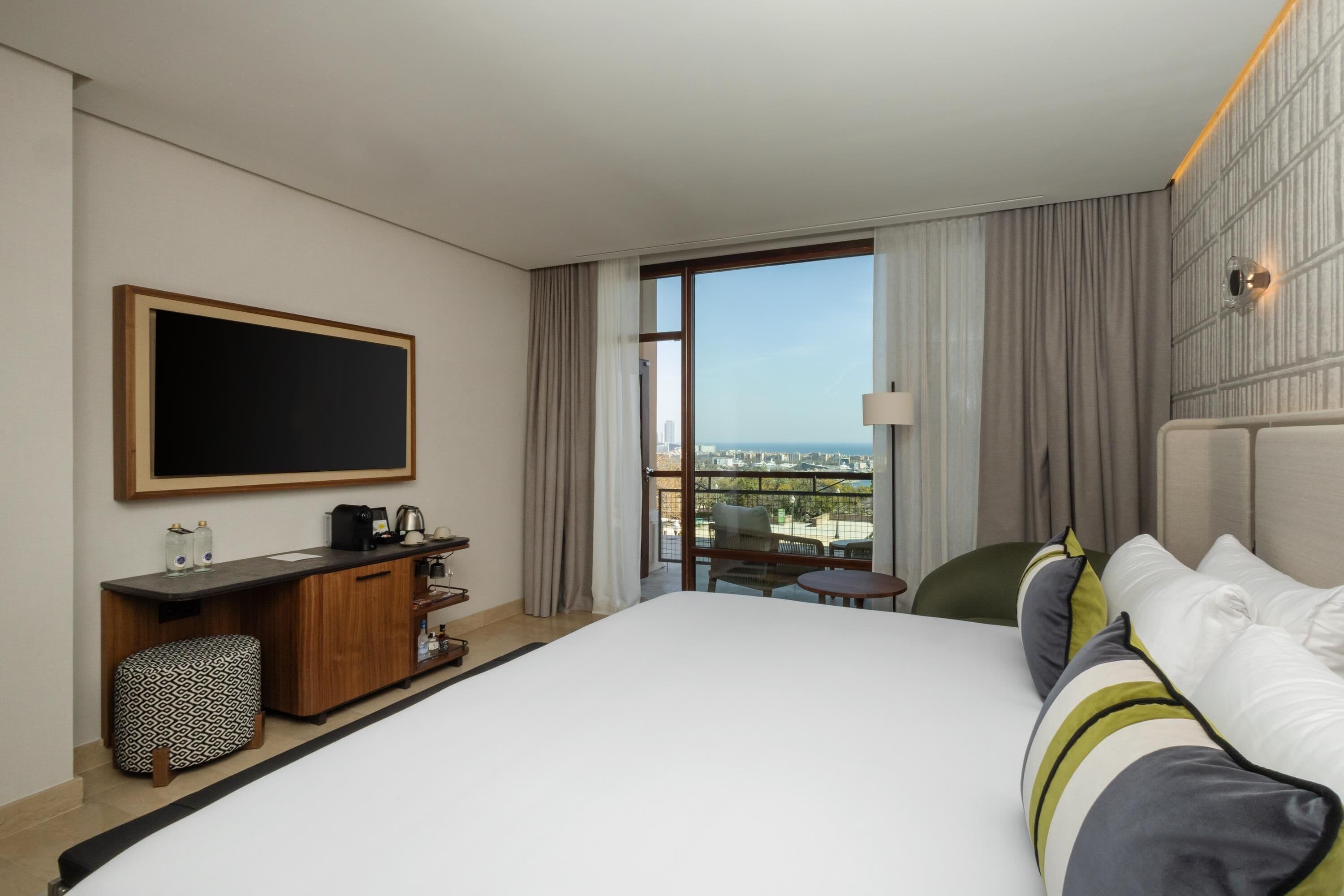 A hotel room with a large bed, a desk with a coffee maker, a TV, and a balcony with a city view.