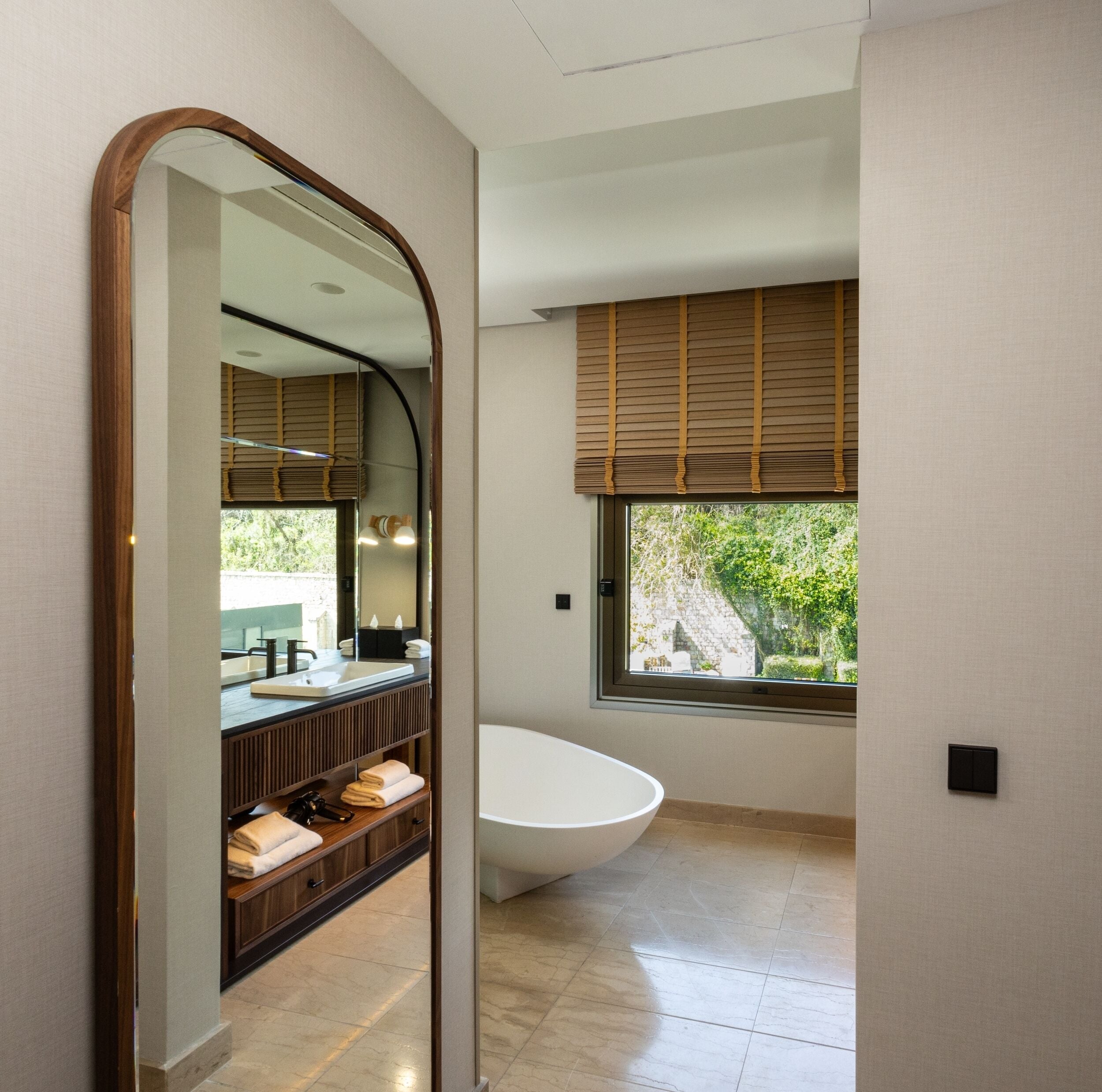 Une salle de bain moderne avec une baignoire sur pieds, un grand miroir et une fenêtre avec des stores.