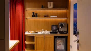 Una kitchenette compacta con cafetera, un refrigerador pequeño y una estantería con un cartel que dice "101MIVING".