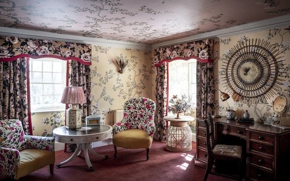 A room with floral wallpaper, a round table with a lamp, a yellow armchair, a wooden cabinet, and a decorative mirror.