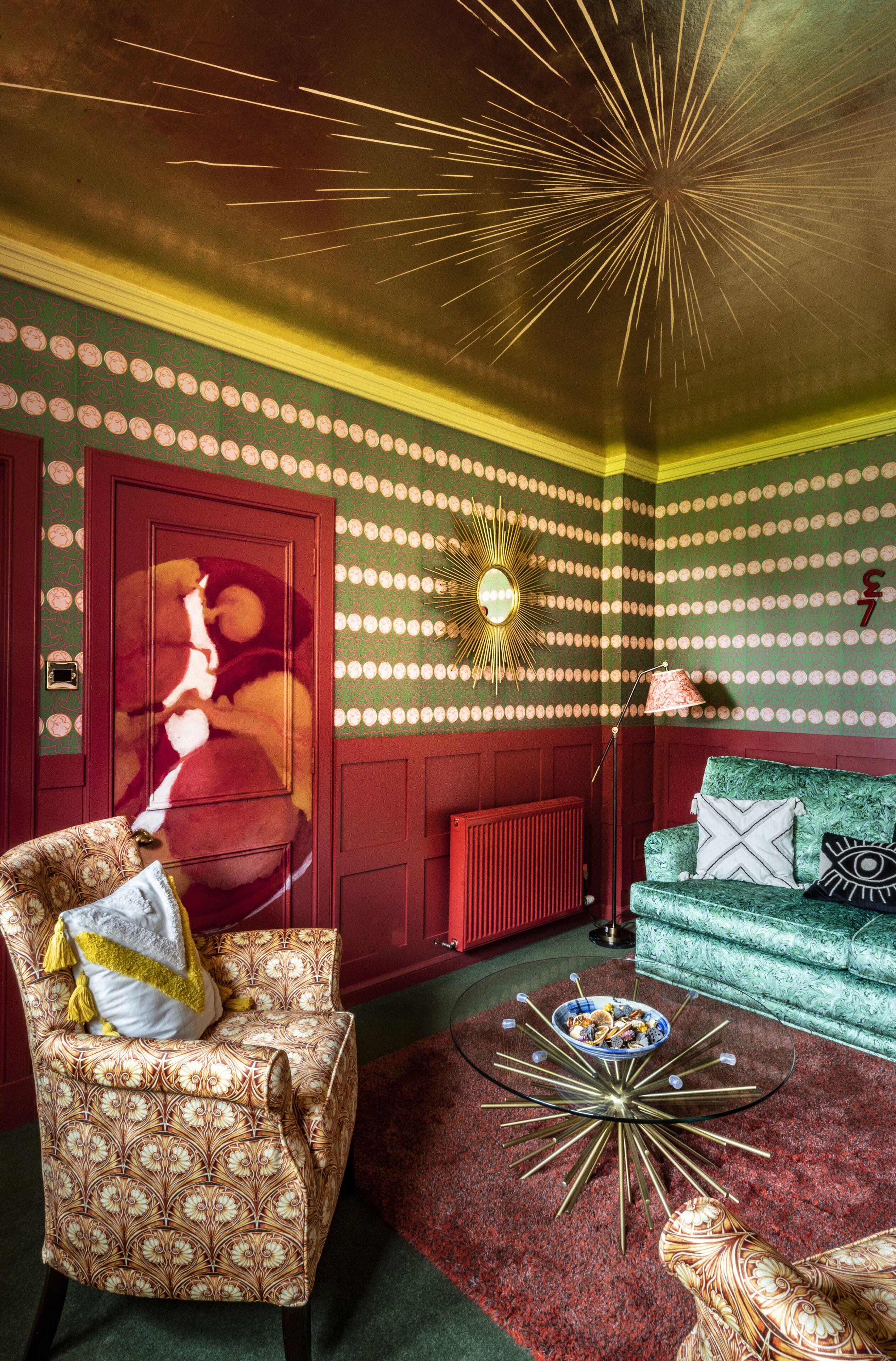 A room with a patterned wall, a red door, a green sofa, a patterned armchair, and a coffee table with a bowl on it.