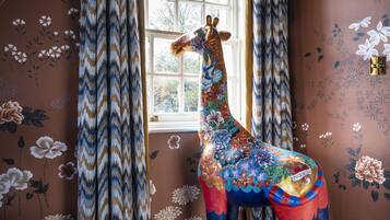 A giraffe statue in a room with patterned wallpaper, a window with curtains, and a carpeted floor.