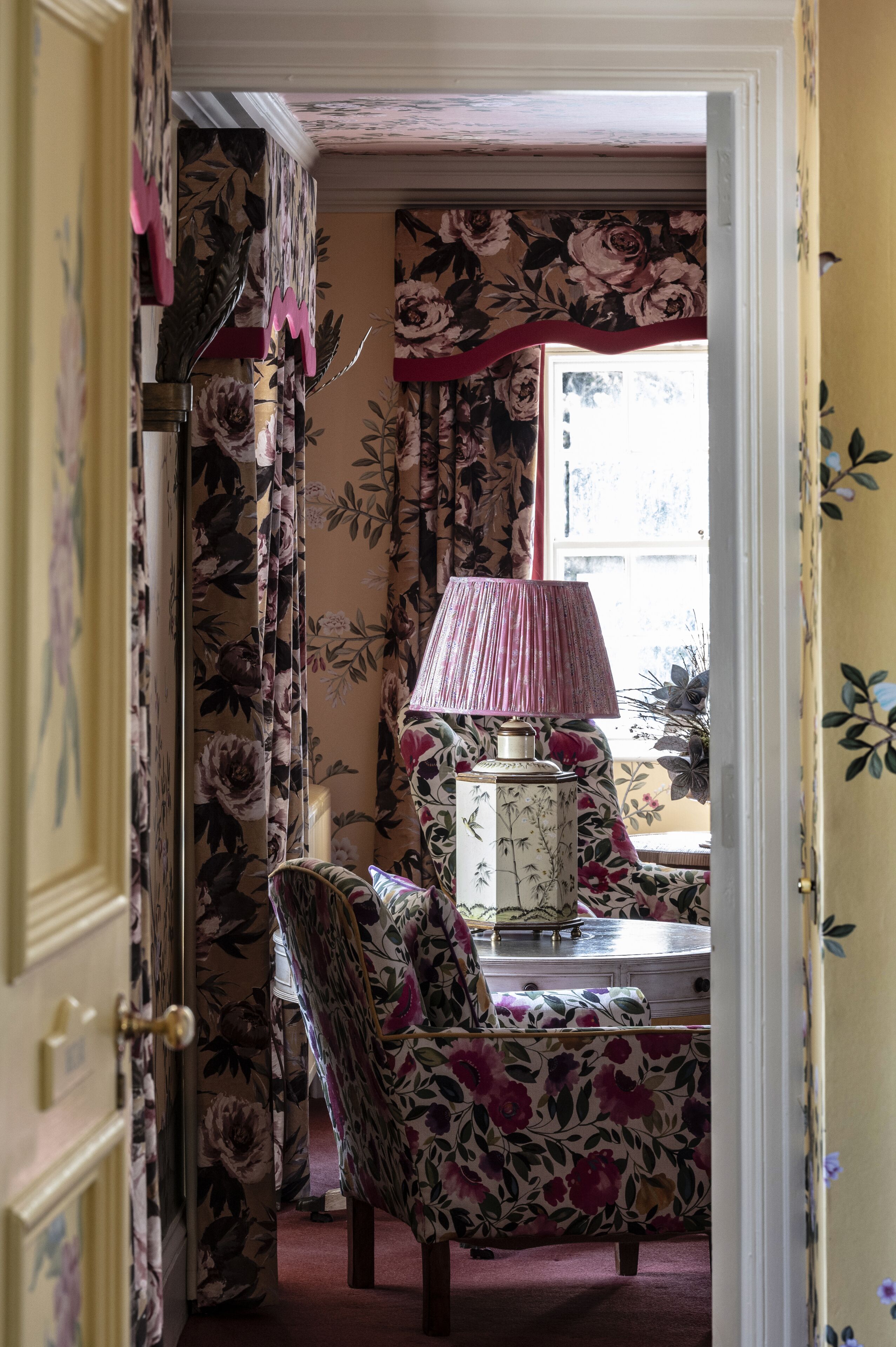 A room with floral wallpaper, a chair with a floral pattern, a table with a lamp, and a window with a view.