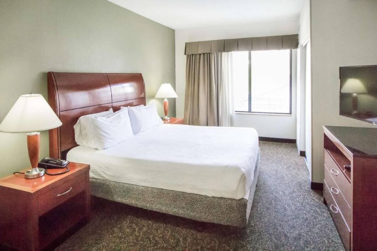 A hotel room with a bed, two bedside tables with lamps, a dresser, a television, and a window with curtains.