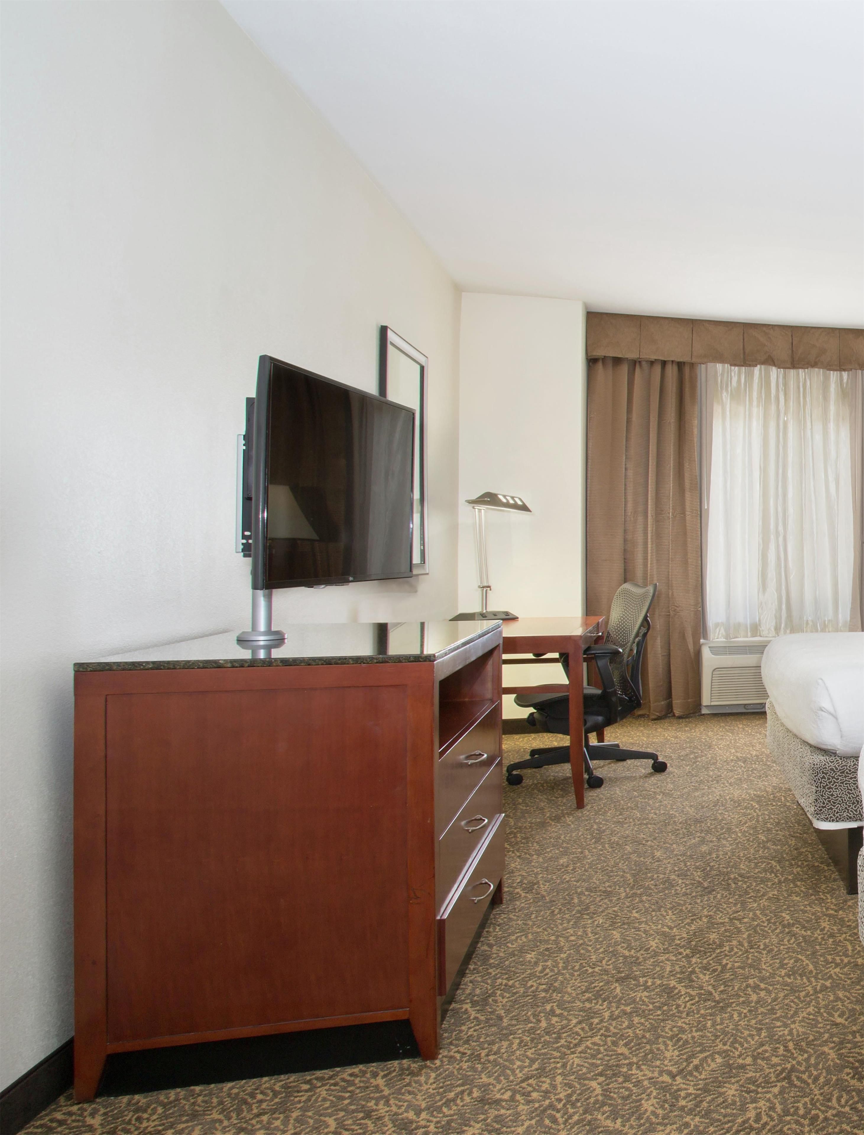 A hotel room with a wooden desk, a television, a chair, a bed, and a window with curtains.