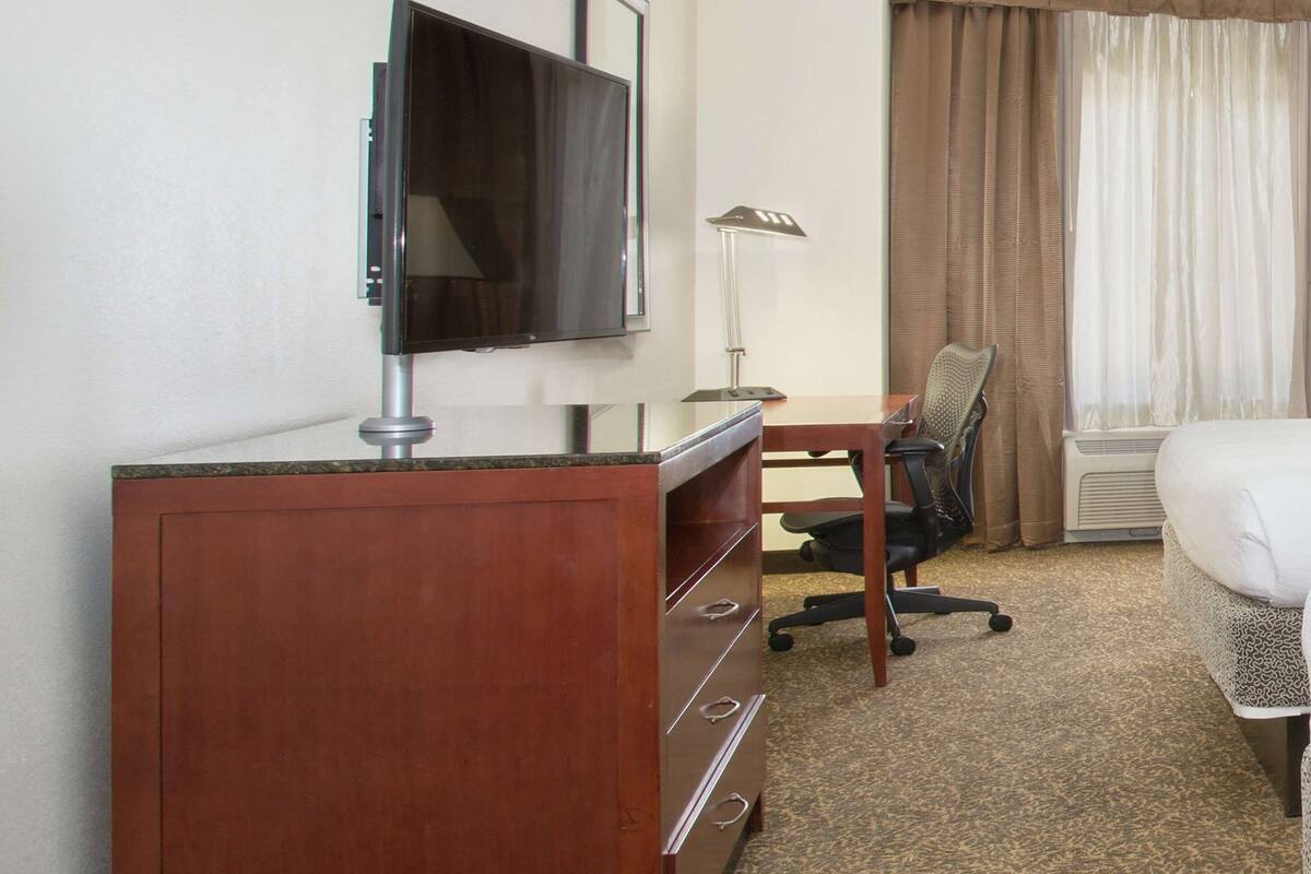 A hotel room with a wooden desk, a television, a chair, a bed, and a window with curtains.