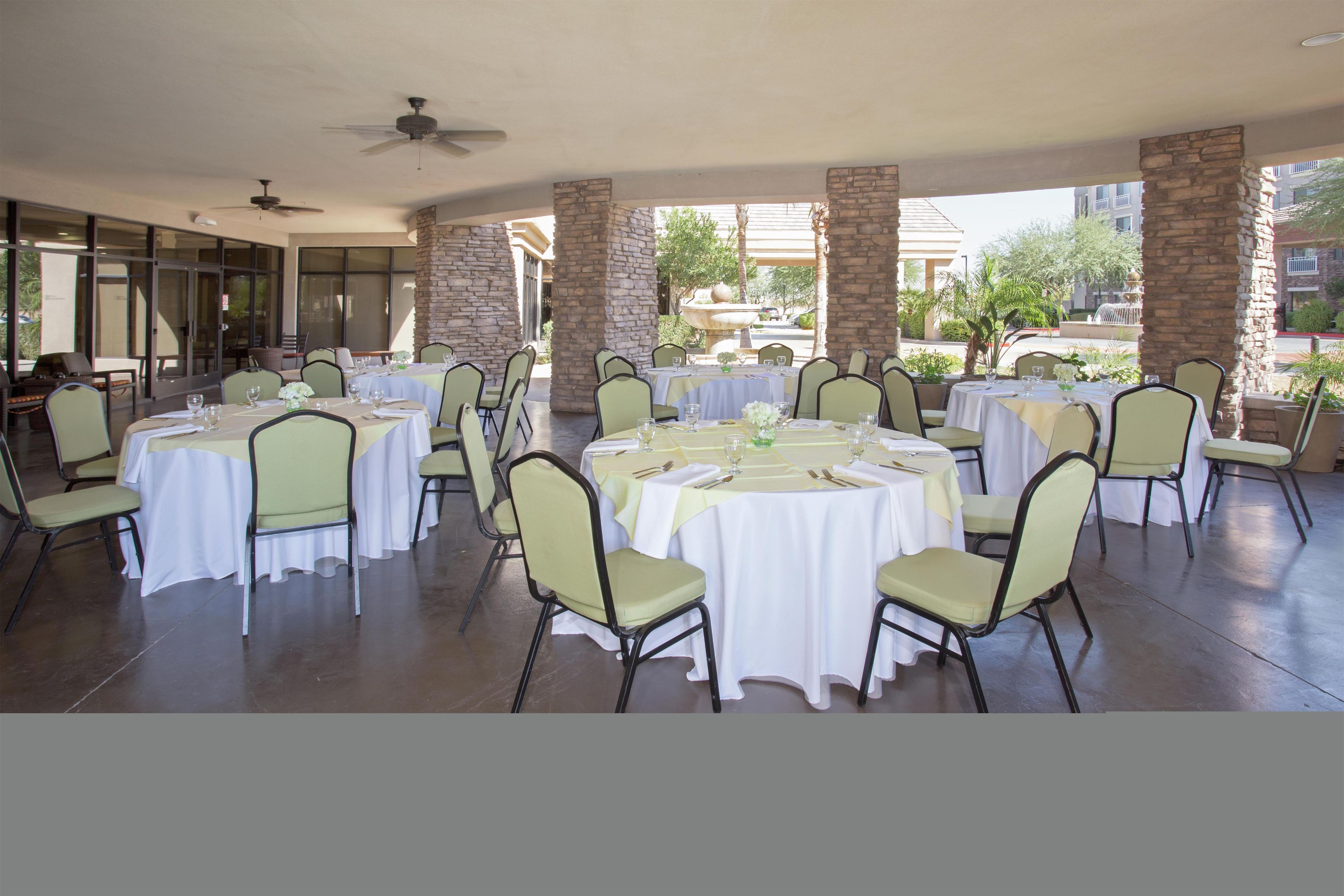 A covered outdoor dining area with round tables and chairs, stone pillars, and a view of buildings and trees.