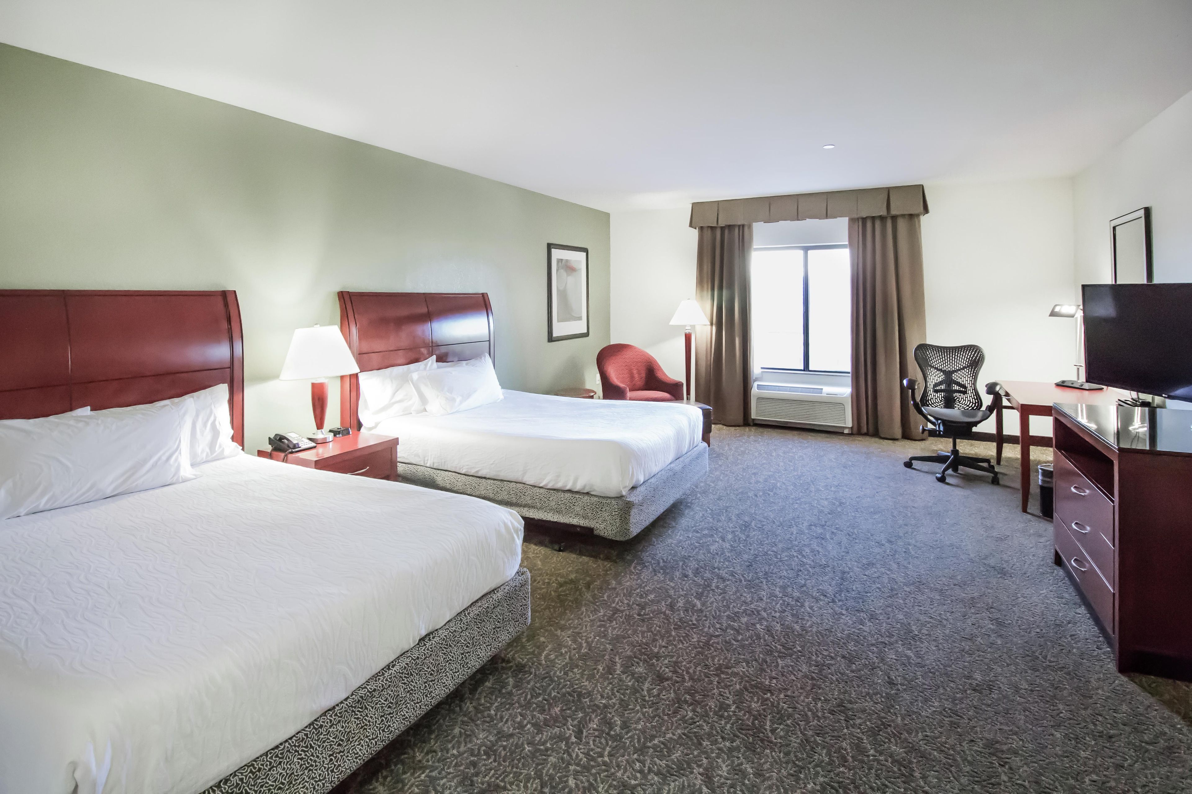 A hotel room with two beds, a desk, a chair, a television, and a window with curtains.