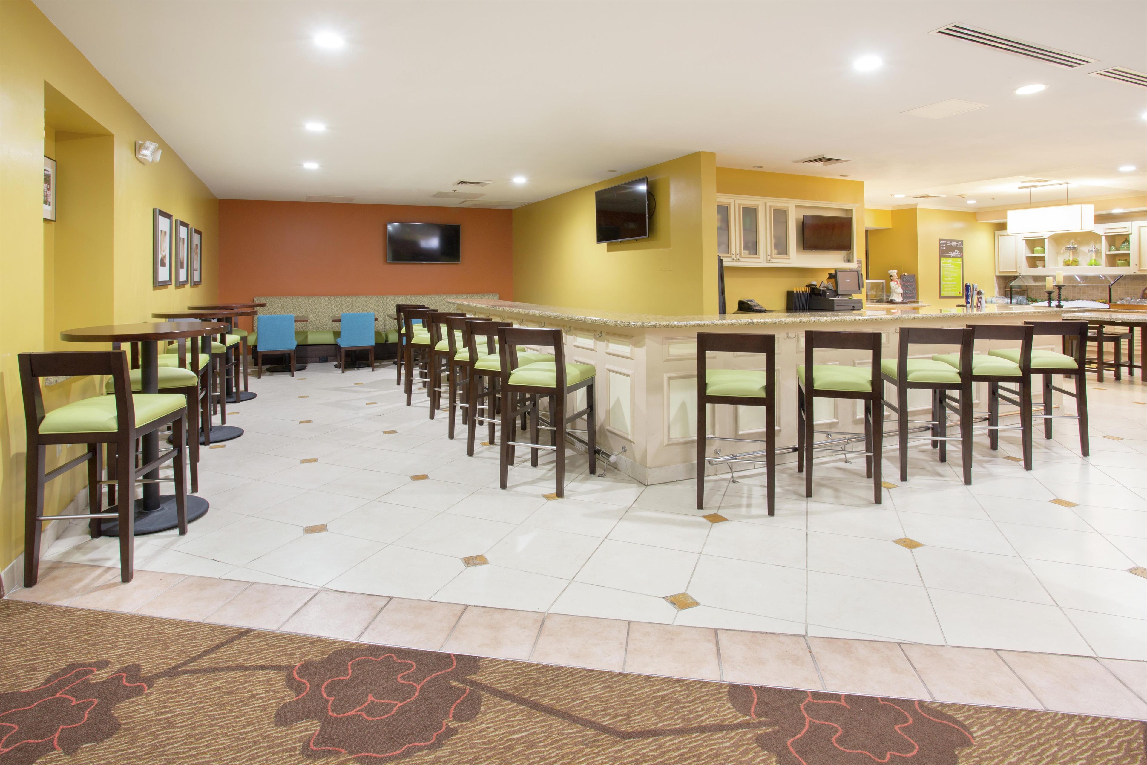 A hotel bar area with multiple seating arrangements, a television, and a kitchen area in the background.