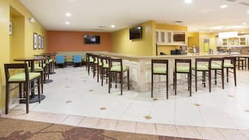 A hotel bar area with multiple seating arrangements, a television, and a kitchen area in the background.