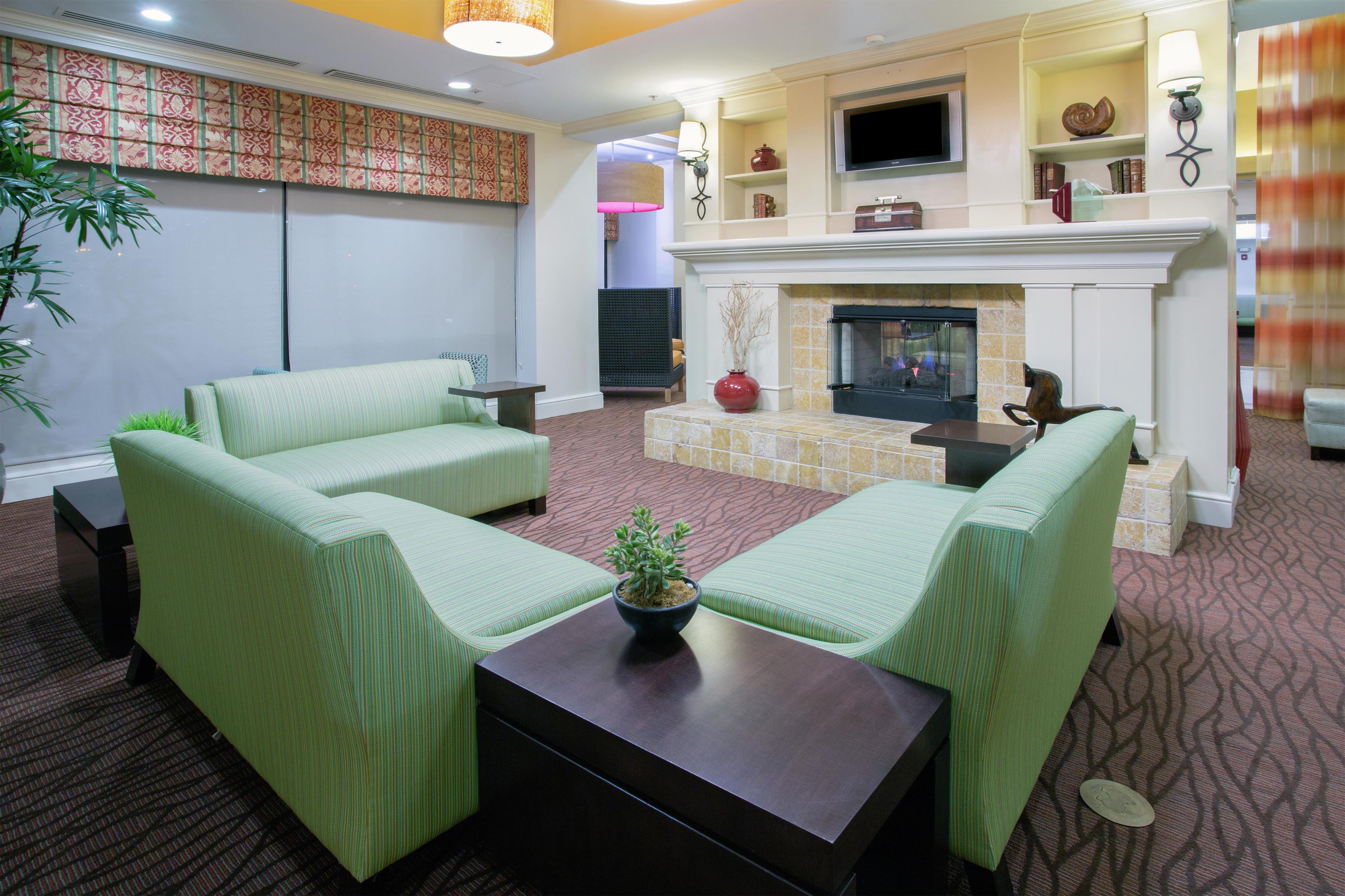 A hotel room with a fireplace, two green sofas, a coffee table, and a large window with blinds.