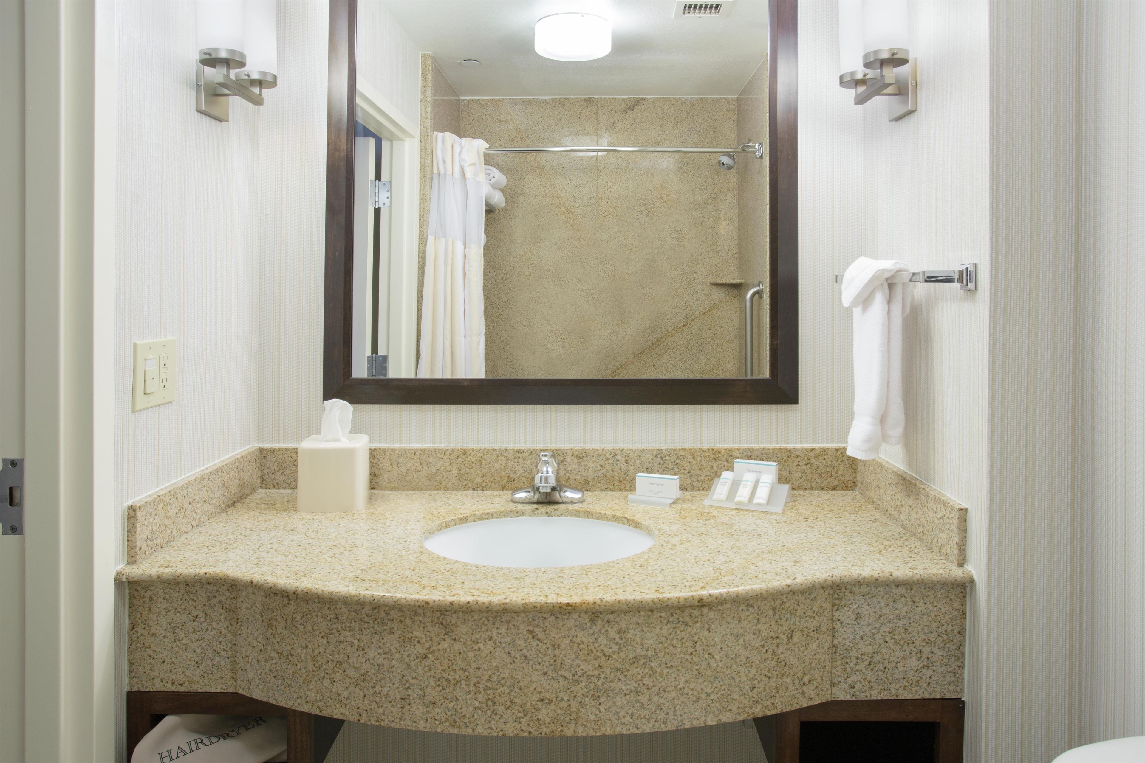 Une salle de bain avec un lavabo doté d’un plan de travail en granit, un miroir, un porte-serviettes et un distributeur de papier toilette.