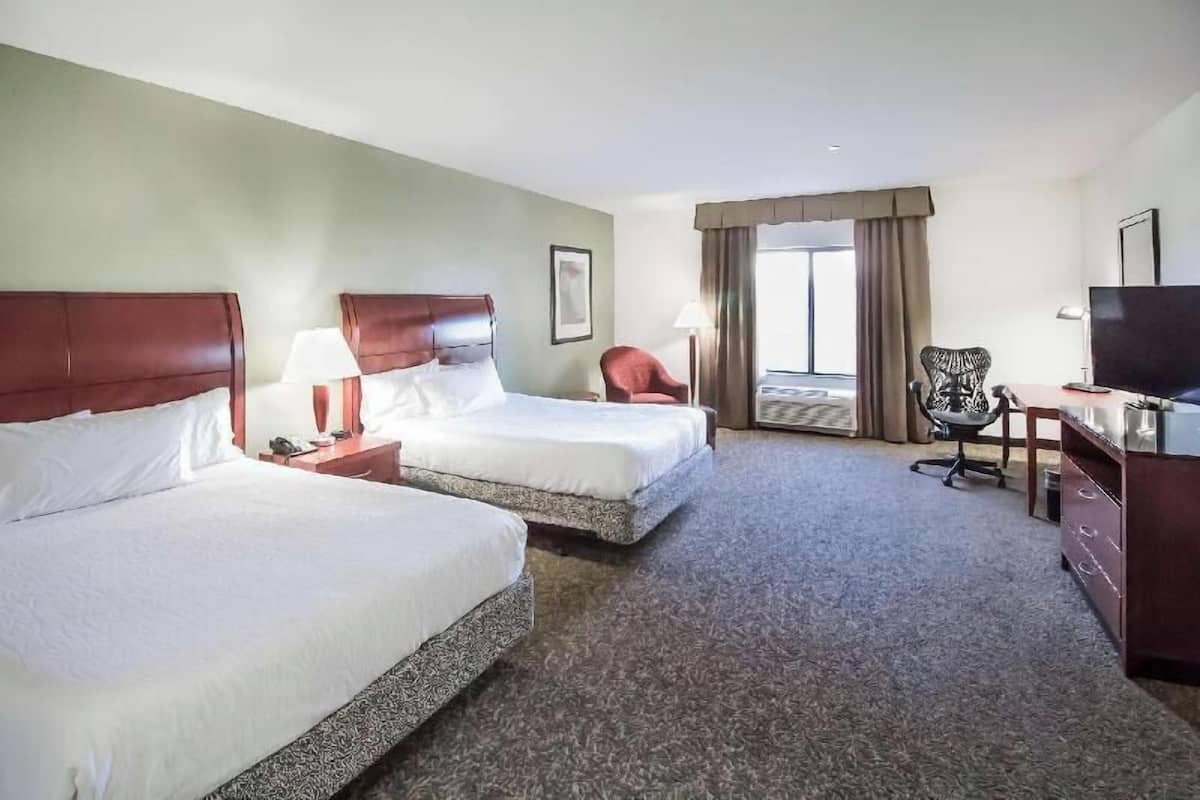 A hotel room with two beds, a desk, a chair, a TV, and a window with curtains.