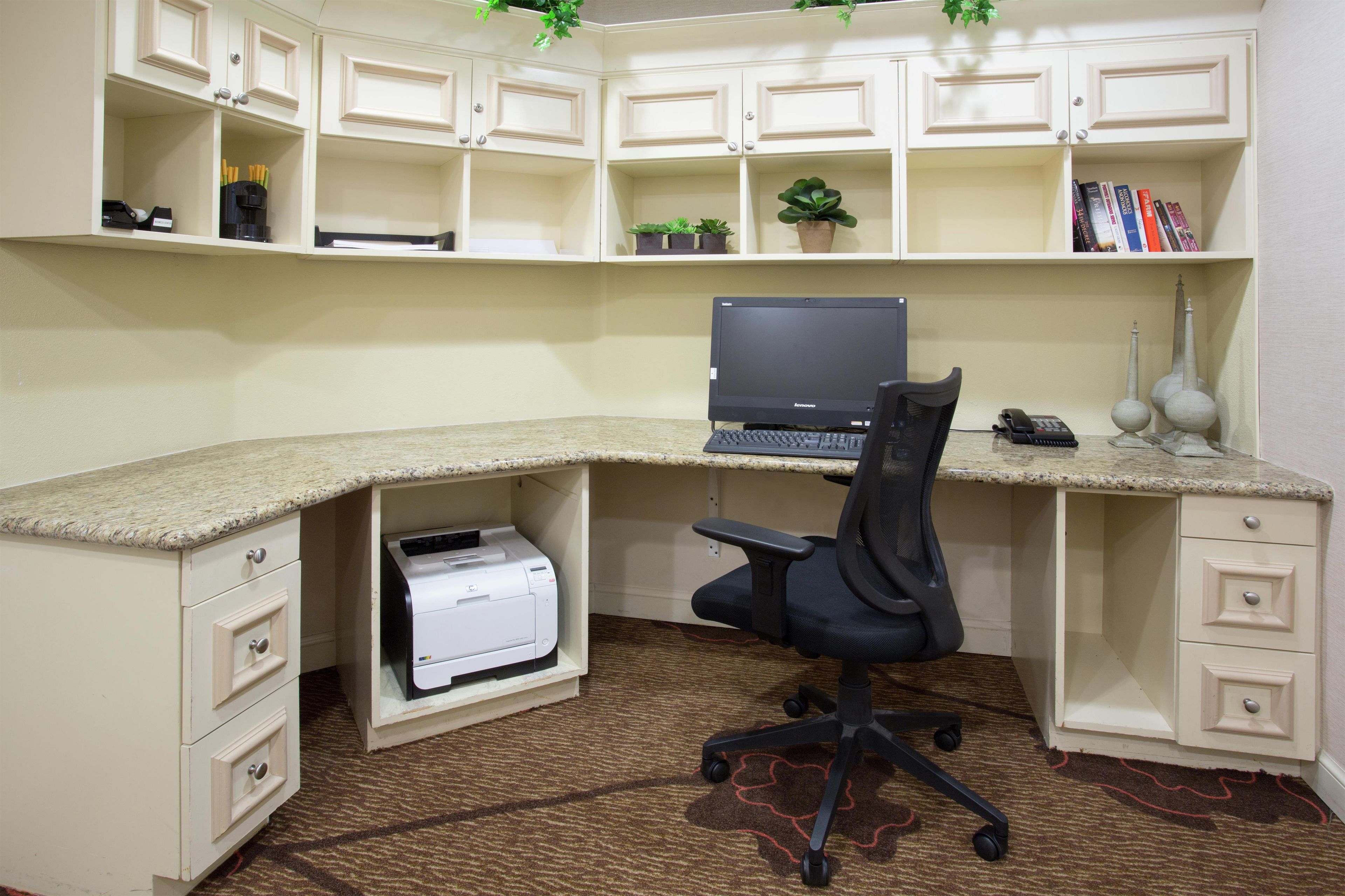 A corner office with a desktop computer, a printer, and a chair. There are shelves above with books and plants.