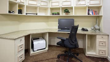 A corner office with a desktop computer, a printer, and a chair. There are shelves above with books and plants.