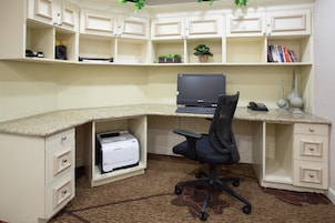 A corner office with a desktop computer, a printer, and a chair. There are shelves above with books and plants.