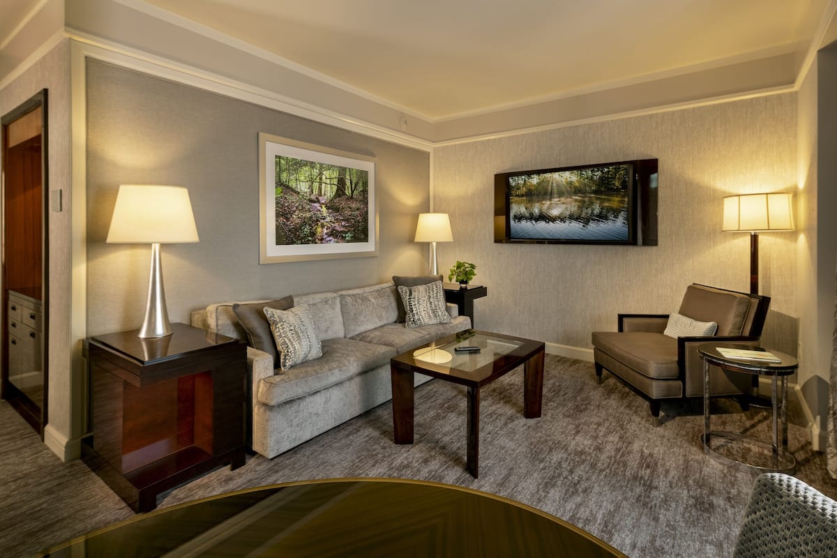A modern living room with a sofa, armchairs, a coffee table, and two lamps. There are two framed pictures on the wall and a flat-screen TV.