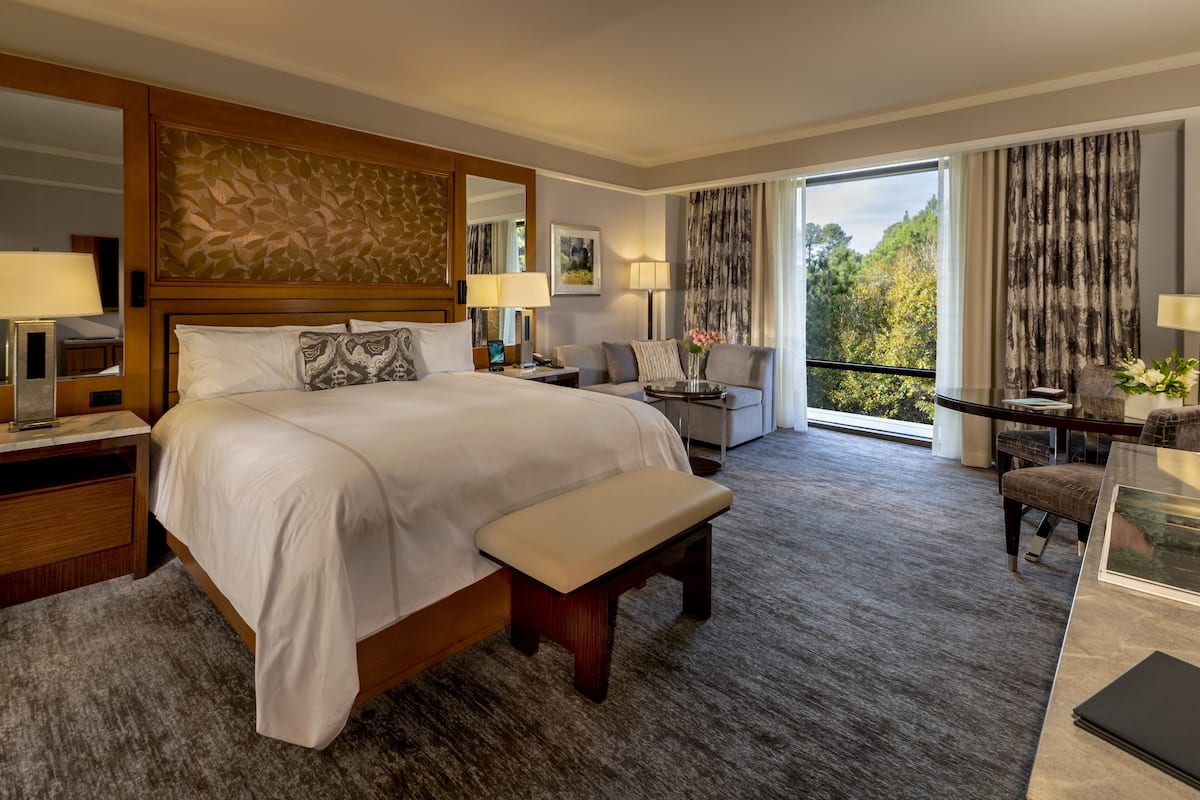 A spacious hotel room with a large bed, a bench, a desk, and a balcony with a view of trees.
