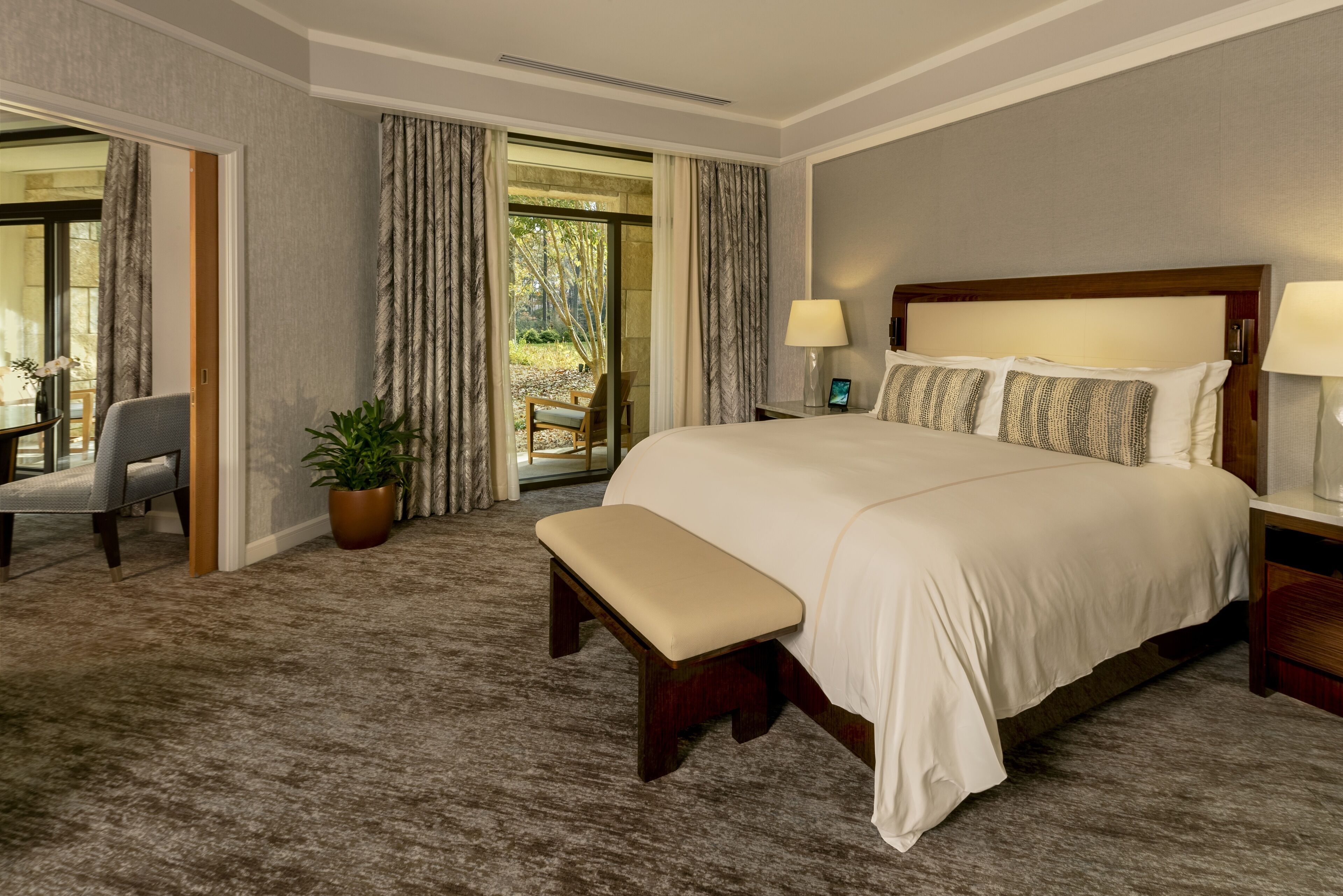 A hotel room with a large bed, a bench, a desk with a lamp, and a view of the outdoors through a glass door.