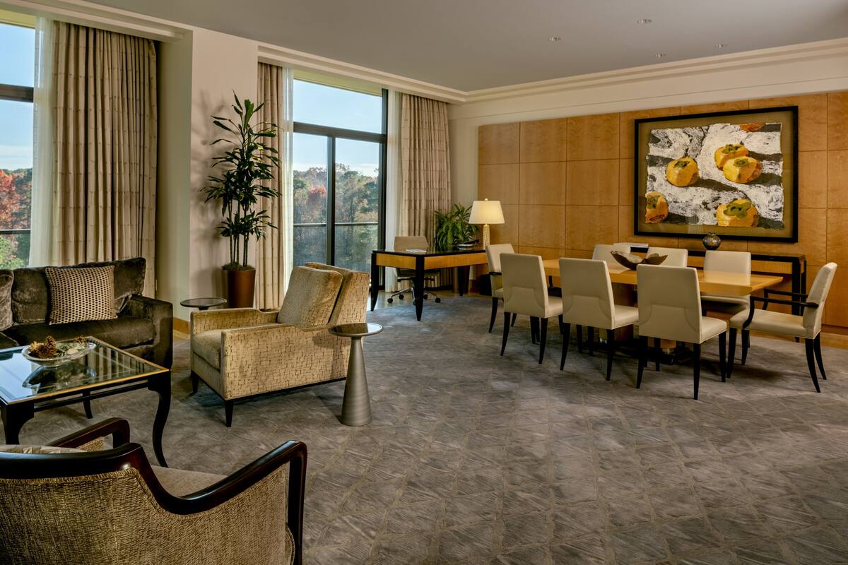 A spacious living area with a sofa, armchairs, a coffee table, a dining table with chairs, and a large window offering a view of trees.