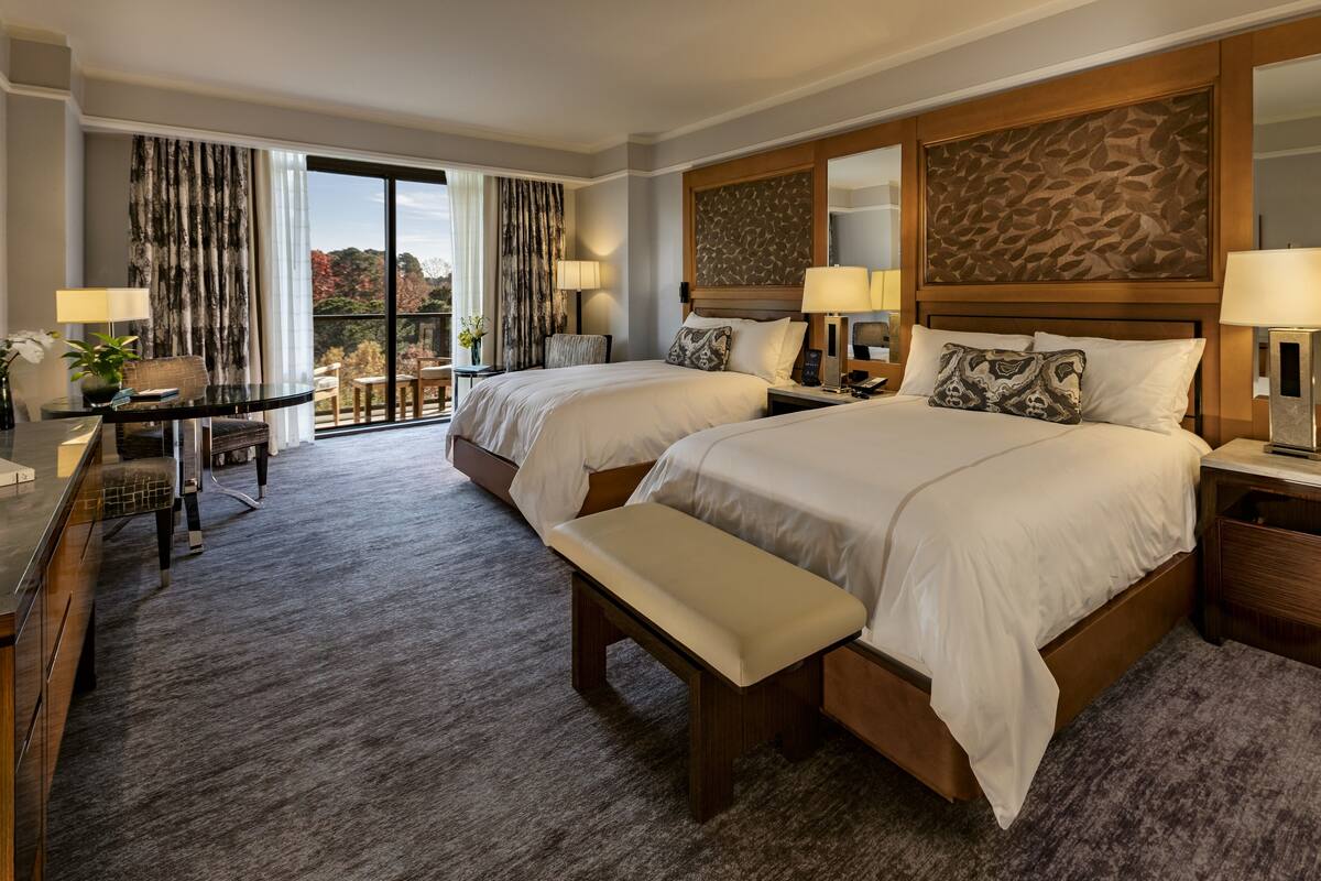 A hotel room with two beds, a desk, a bench, and a view of the outdoors.
