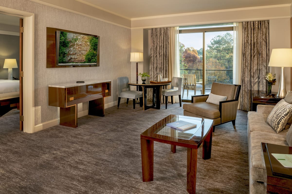 A hotel room with a large bed, a desk, a sofa, and a balcony with a view of trees.
