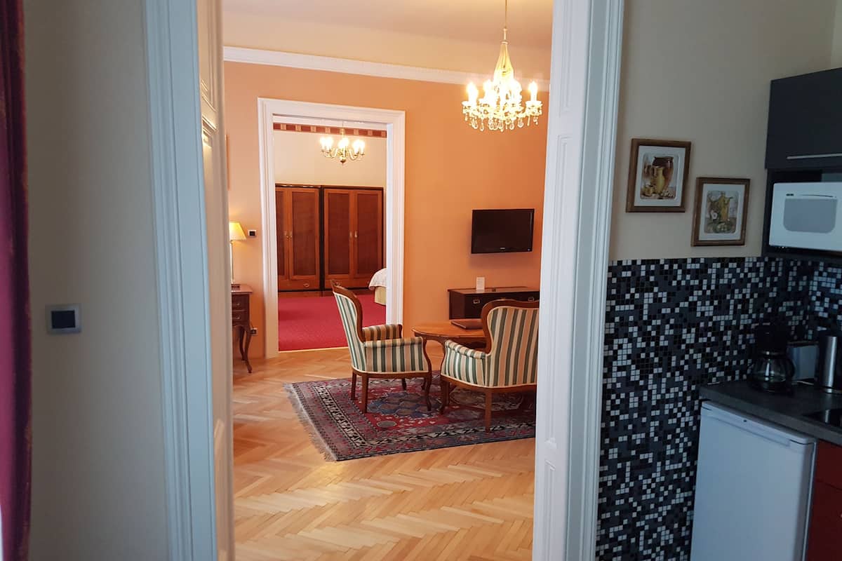 A hotel room with a wooden herringbone floor, a dining area with striped chairs, a chandelier, a TV, and a kitchenette with a microwave.
