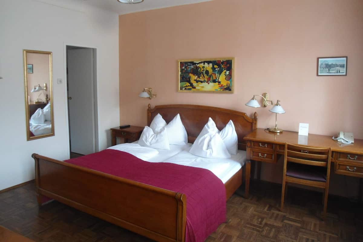 A hotel room with a wooden bed, a desk, a chair, a mirror, and a painting on the wall.