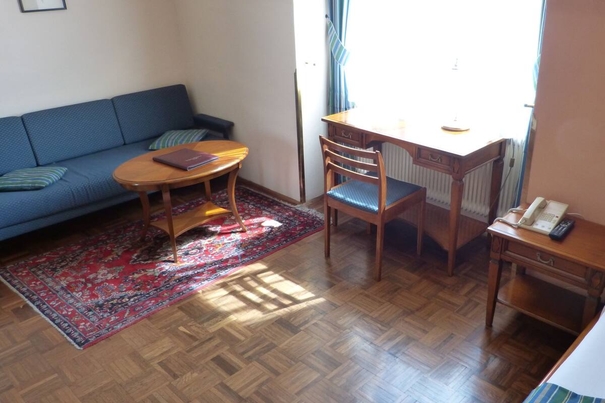 A room with a wooden desk, a chair, a sofa, a small table, a telephone, and a bed.