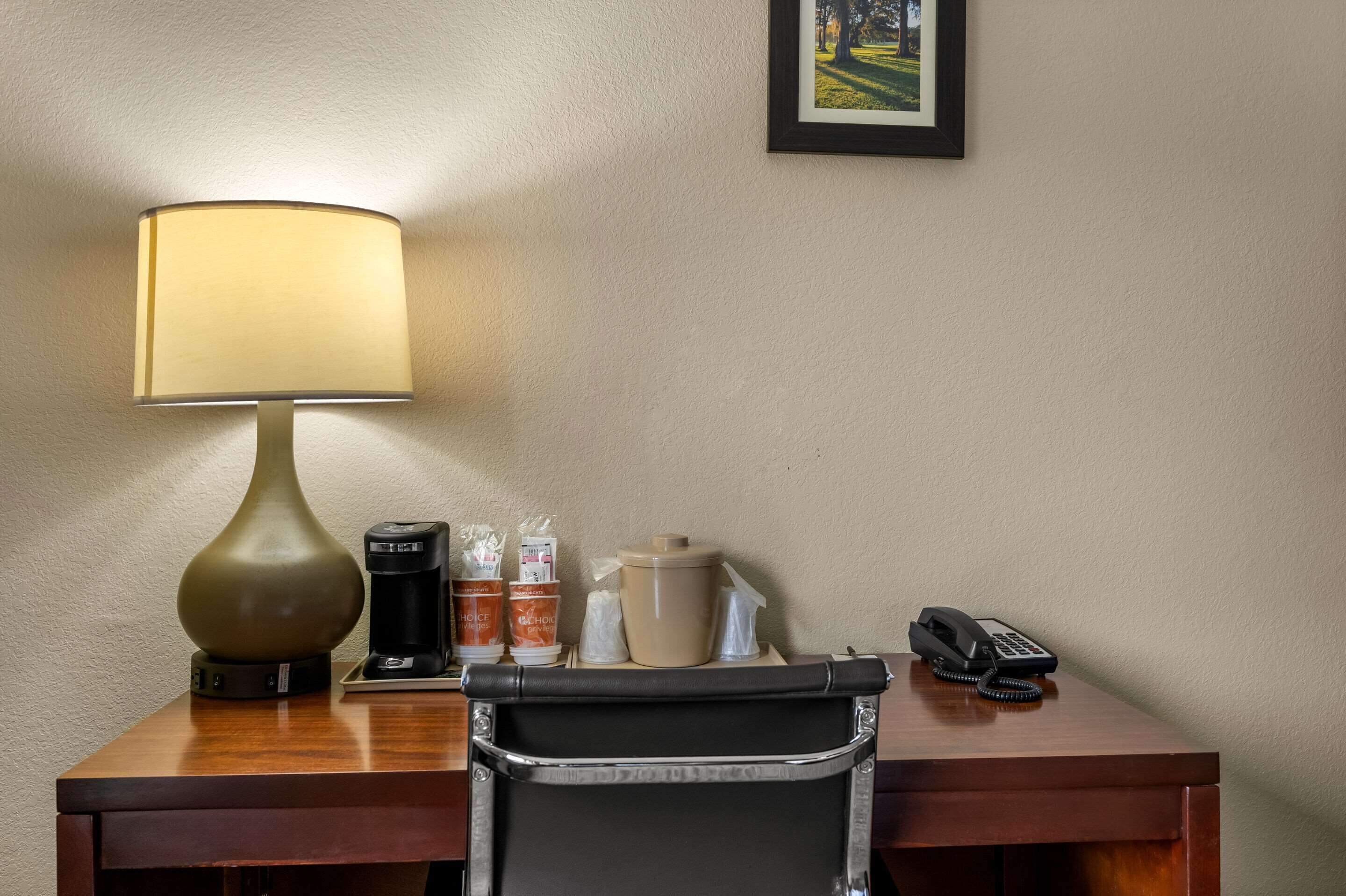 Hotelkamer met een bureau, een lamp, een koffiezetapparaat, een telefoon en een ingelijst kunstwerk aan de muur.