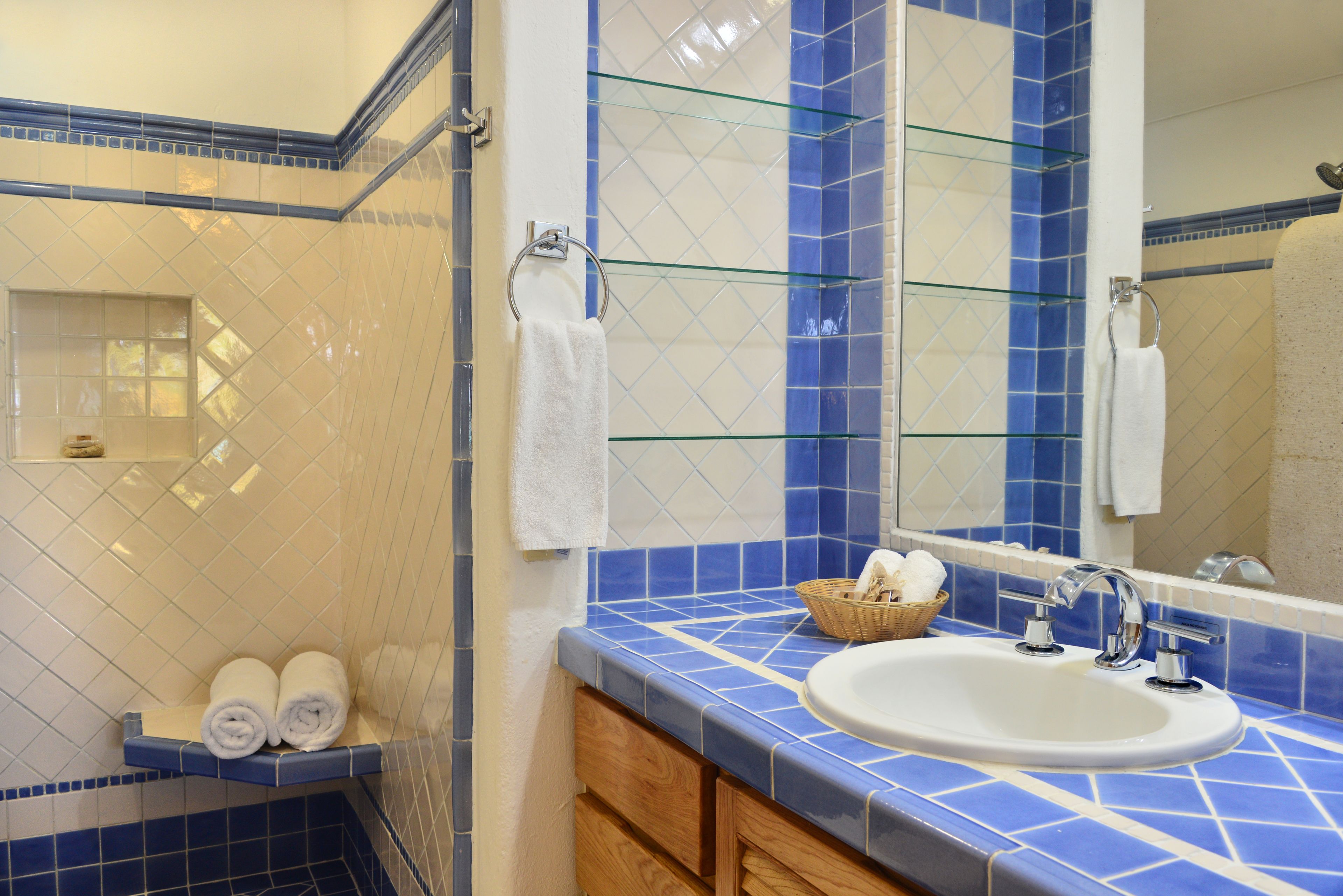 A bathroom with a blue-tiled wall, a white sink, and a glass shelf holding towels and a basket.