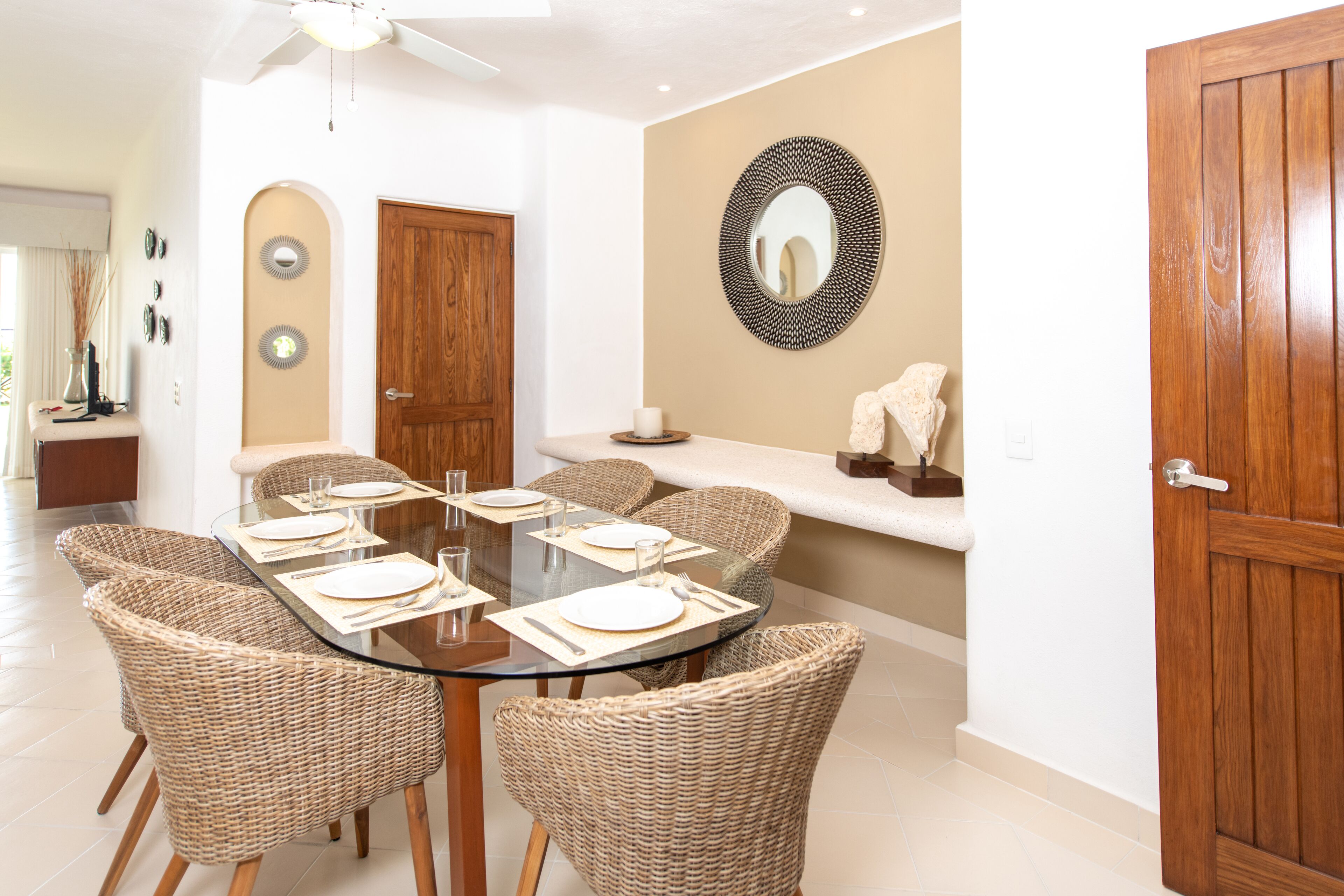 A dining area with a round glass table surrounded by wicker chairs, a wooden door, and a wall-mounted mirror.