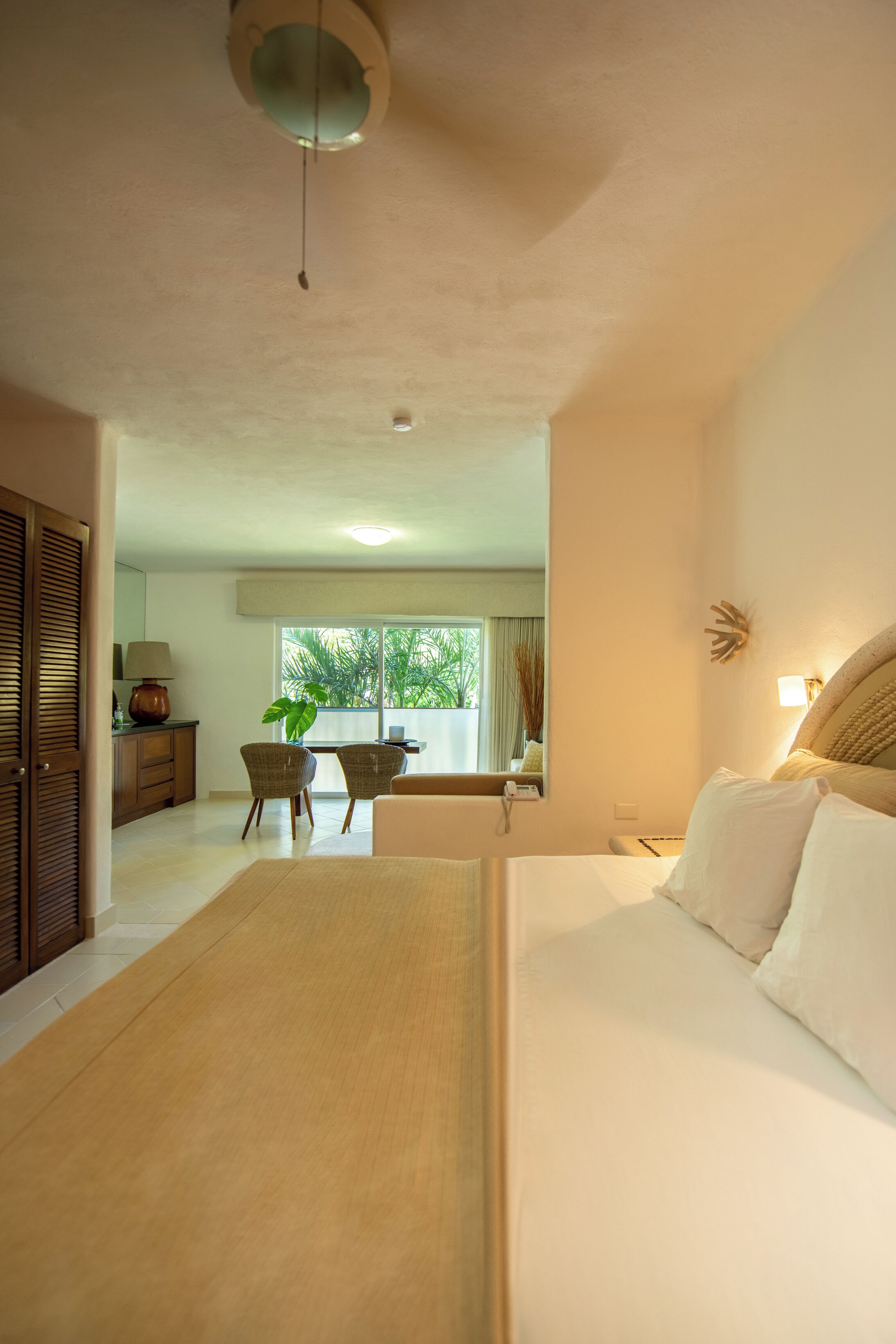 A hotel room with a bed, a desk, chairs, a plant, and a view of the outdoors.