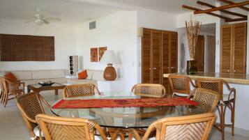 A dining area with a glass table surrounded by wicker chairs, a sofa with orange cushions, and a wall-mounted lamp.