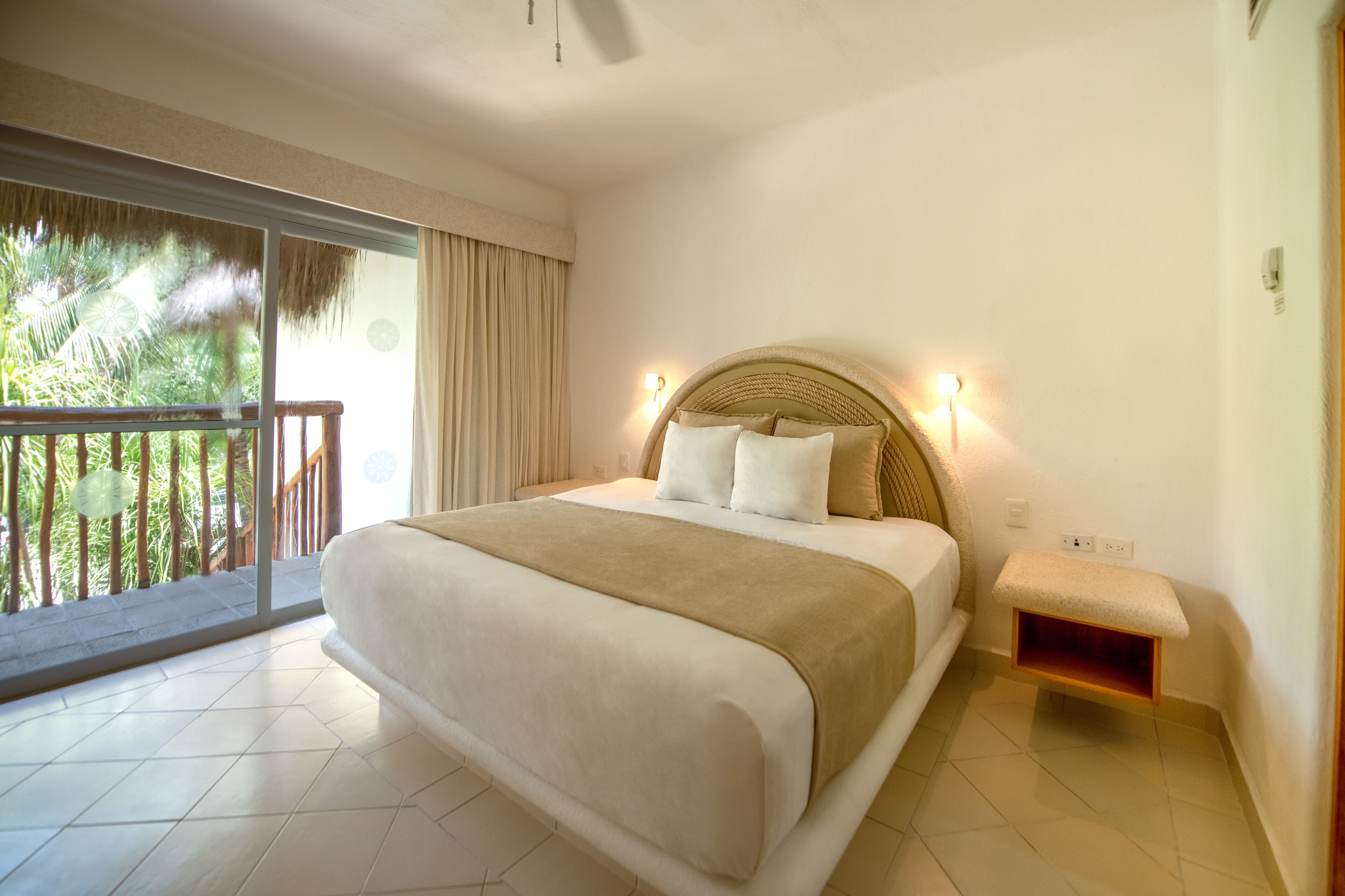 A large bed with a headboard, a small bench, and a balcony with a view of greenery.