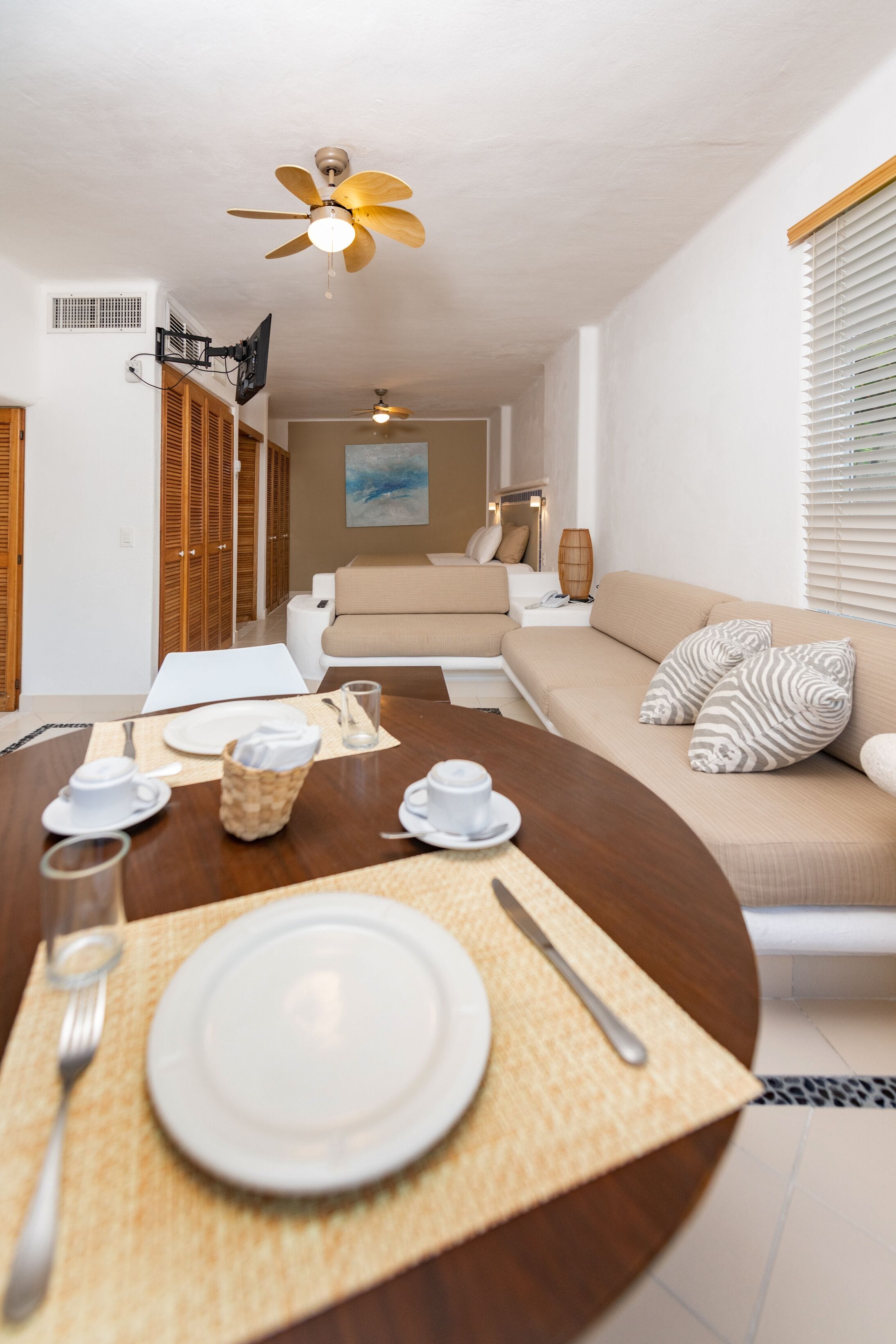 A dining area with a round table set for two, a sofa with patterned cushions, a ceiling fan, and a television mounted on the wall.