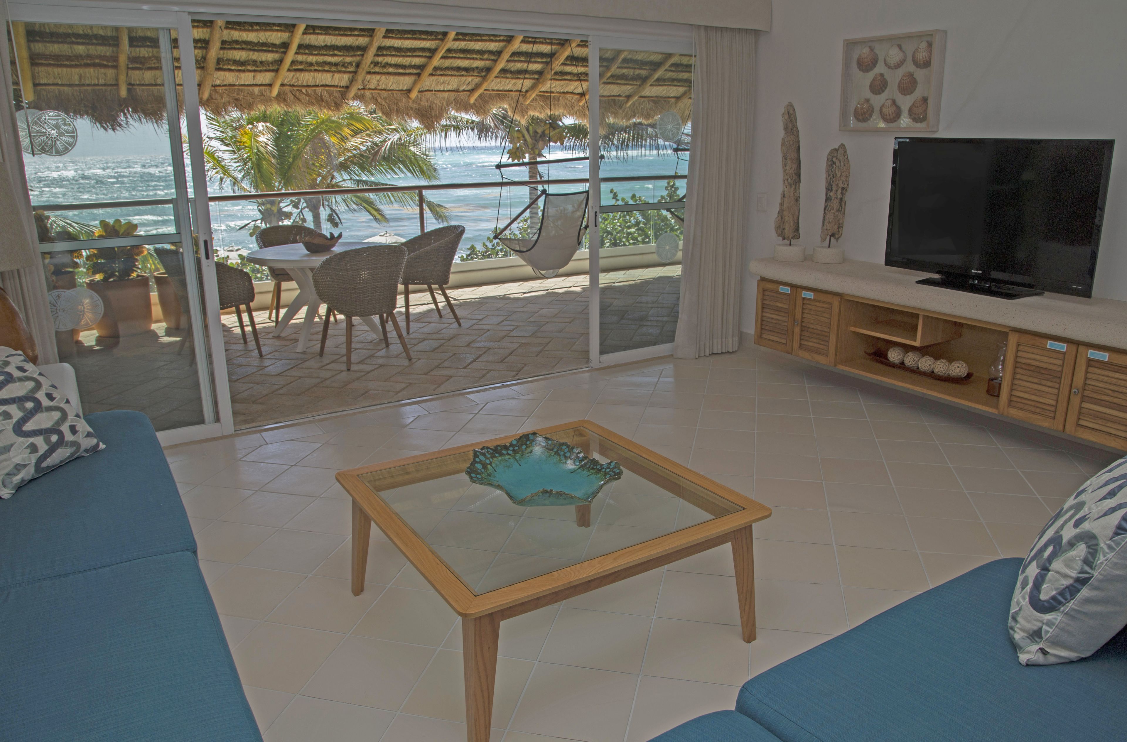 Un salón con mesa de vidrio, sofá azul, televisor y vista al océano.