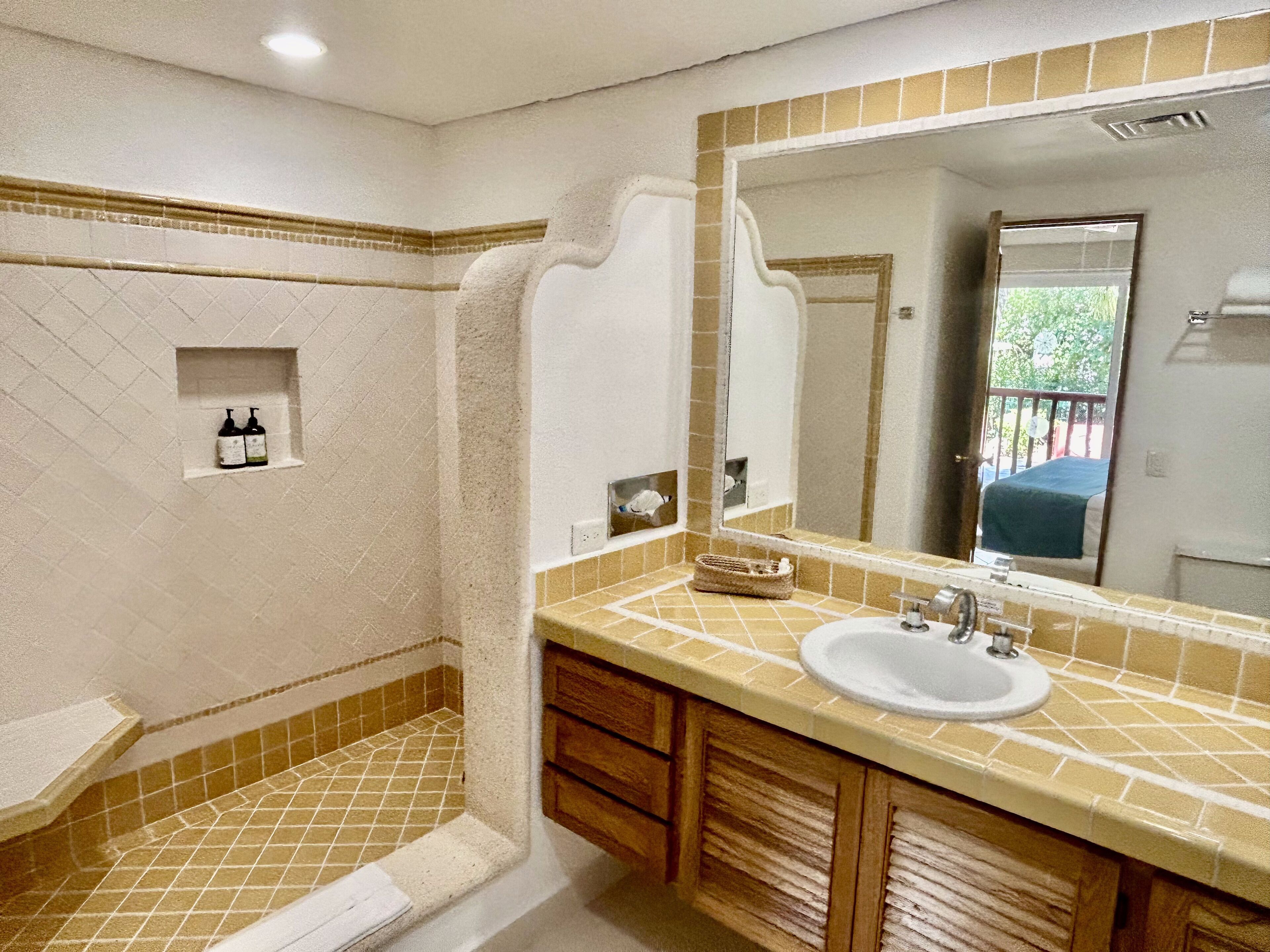 A bathroom with a shower, sink, and mirror. There are toiletries in a shelf and a towel rack.