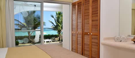 A bedroom with a bed, a wooden wardrobe, a bathroom with a sink and towel rack, and a view of the beach and palm trees through a glass door.