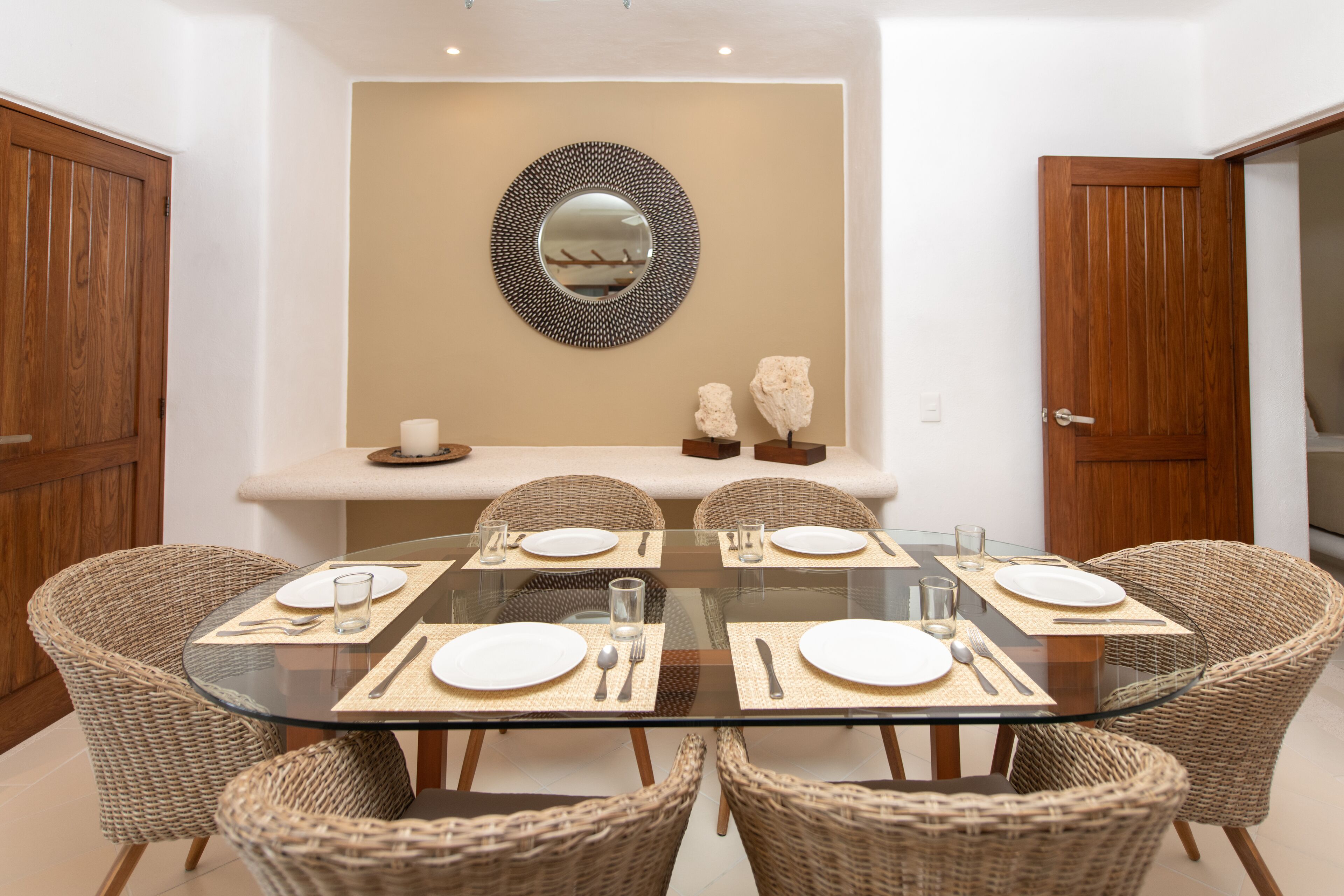 A dining area with a glass table surrounded by wicker chairs, a decorative mirror on the wall, and a wooden door on the right.