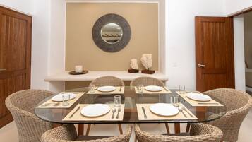 A dining area with a glass table surrounded by wicker chairs, a decorative mirror on the wall, and a wooden door on the right.