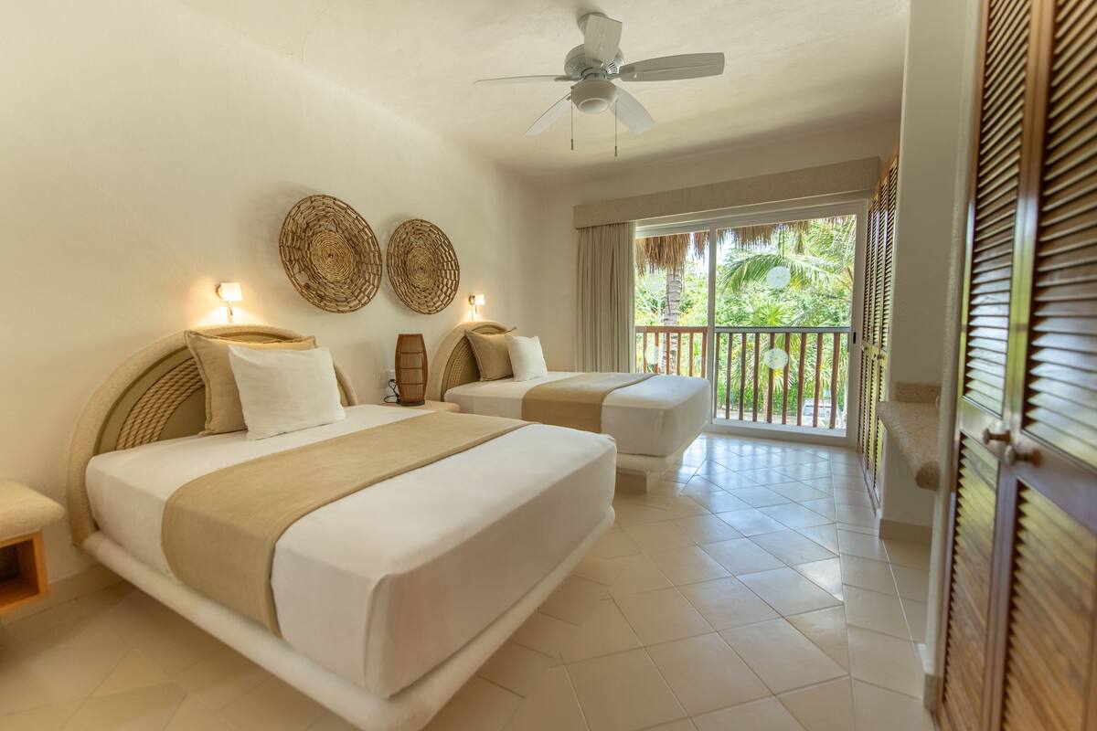 A hotel room with two beds, a ceiling fan, woven wall decor, and a balcony with a view of greenery.